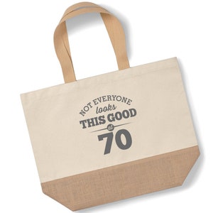 Puede incluir: Una bolsa de lona beige con ribete de yute marr&oacute;n. La bolsa tiene asas marrones y presenta el texto "Not everyone looks this good at 70".