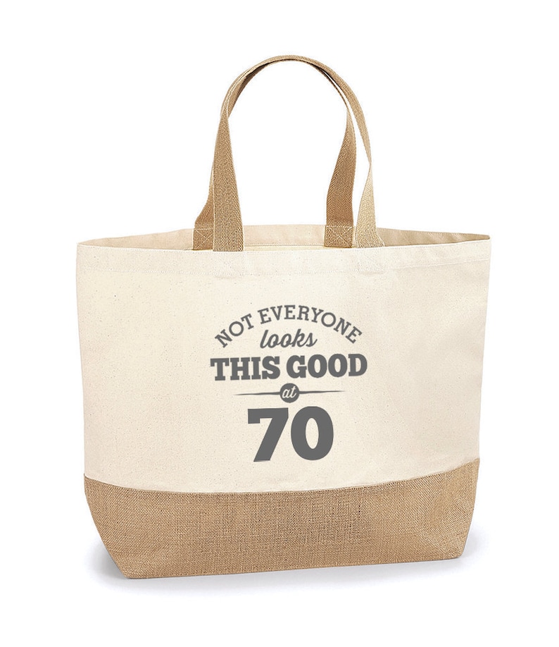 Puede incluir: Una bolsa de lona beige con ribete de yute marr&oacute;n. La bolsa tiene una impresi&oacute;n gris que dice "Not everyone looks this good at 70".