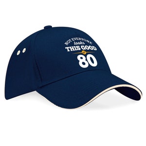 May include: A navy blue baseball cap with a white brim and a white inscription that reads "Not everyone looks this good at 80".