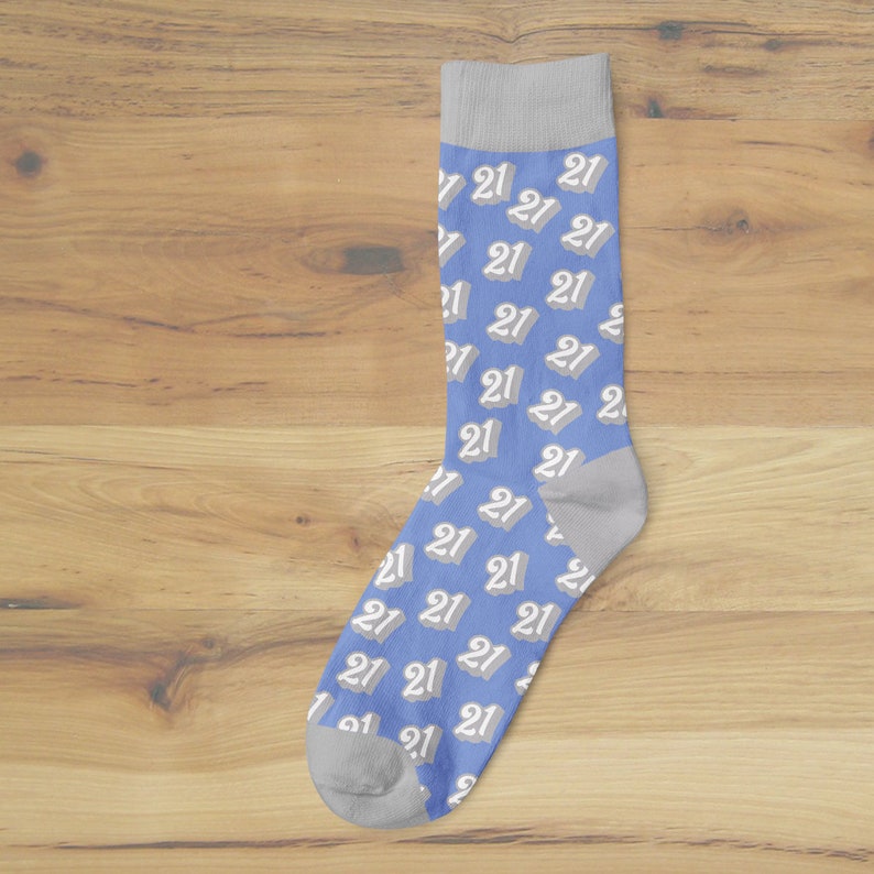 May include: A blue sock with a pattern of the number "21" in white and gray. The sock has a gray heel and toe, and a gray cuff. The sock is displayed on a wooden surface.