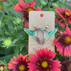 May include: Hummingbird earrings with green and white wings, red and brown bodies, and gold-colored accents. The earrings are displayed on a brown card with the text "KE&JUL HANDMADE JEWELRY". The background features red and yellow flowers.