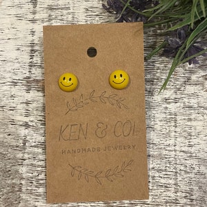 Op de afbeelding: Gele smiley-oorbellen op een bruine kaart. De oorbellen hebben een zwart lachend gezicht. De kaart is voorzien van de tekst "KEN & COL HANDMADE JEWELRY". De kaart heeft een klein gaatje aan de bovenkant.