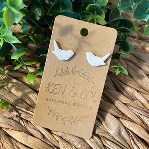 May include: White bird-shaped stud earrings on a brown card with the text "KEN & COL HANDMADE JEWELRY". The earrings have a speckled texture. The card is displayed on a woven, tan surface with green foliage in the background.