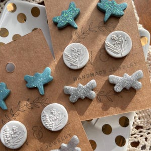 May include: Handmade jewelry earrings on display. The earrings include dragonfly and floral designs in shades of blue and silver. The earrings are displayed on brown cards with the words "HANDMADE JEWELRY" printed on them.