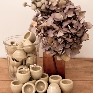 Op de afbeelding: Een verzameling kleine, lichtgrijze keramische potten, sommige gestapeld op elkaar. De potten zijn gerangschikt op een houten oppervlak. Op de achtergrond staat een vaas met gedroogde bloemen.