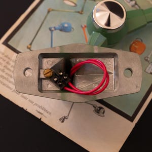 May include: Close-up of a silver electrical component with two red wires connected to a black connector. The component is mounted on a green and silver base.