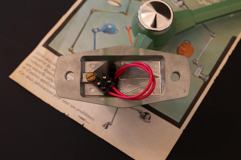 May include: Close-up of a silver electrical component with two red wires connected to a black connector. The component is mounted on a green and silver base.