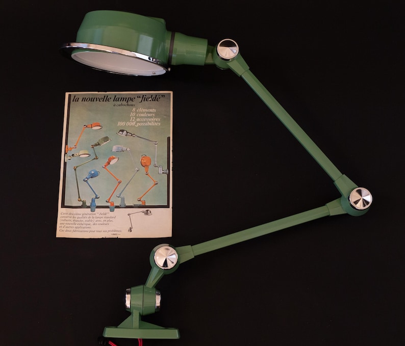 May include: A green metal desk lamp with a silver reflector and a clamp base. The lamp has two adjustable arms and a silver knob on each joint. The lamp is shown on a black background with a brochure for the lamp in the background. The brochure is in French and features the text "la nouvelle lampe "fielde"" and "100 000 possibilit&eacute;s".