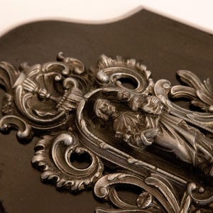 May include: A silver-toned religious plaque with an ornate design featuring a figure of a saint holding a child. The plaque is mounted on a dark wood background.