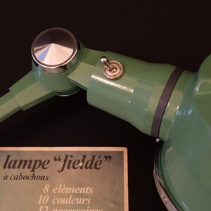 May include: A green metal desk lamp with a silver knob and a toggle switch. The lamp has a black cord and a white label that reads "nouvelle lampe "fielde" &agrave; cabochons 8 &eacute;l&eacute;ments 10 couleurs 12 accessoires".