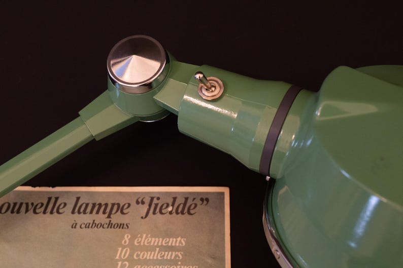 May include: A green metal desk lamp with a silver knob and a toggle switch. The lamp has a black cord and a white label that reads "nouvelle lampe "fielde" &agrave; cabochons 8 &eacute;l&eacute;ments 10 couleurs 12 accessoires".
