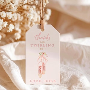 May include: A light pink gift tag with the text "thanks for twirling by!" and "love, sola". The tag features a watercolor illustration of ballet shoes with pink ribbons and floral accents. A brown twine is attached to the tag.