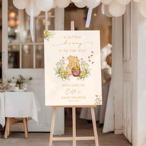 May include: A wooden easel displays a baby shower sign with Winnie the Pooh imagery. The sign reads "A Little Hunny Is On The Way!" with a Pooh illustration and "Welcome to Ella's Baby Shower." White balloons and floral accents decorate the scene.