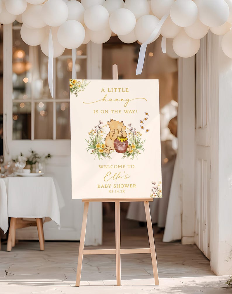 May include: A wooden easel displays a baby shower sign with Winnie the Pooh imagery. The sign reads "A Little Hunny Is On The Way!" with a Pooh illustration and "Welcome to Ella's Baby Shower." White balloons and floral accents decorate the scene.
