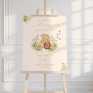 May include: A baby shower sign featuring a Winnie the Pooh illustration with the text "A Little Hunny Is On The Way!" and "Welcome to Ella's Baby Shower 03.14.2X". The sign is on a wooden easel, with floral accents.
