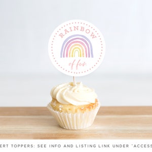 May include: A white cupcake with vanilla frosting topped with a round, white paper cupcake topper that says "RAINBOW of fun" in pink lettering. The topper features a watercolor rainbow with purple, pink, yellow, and orange colors.