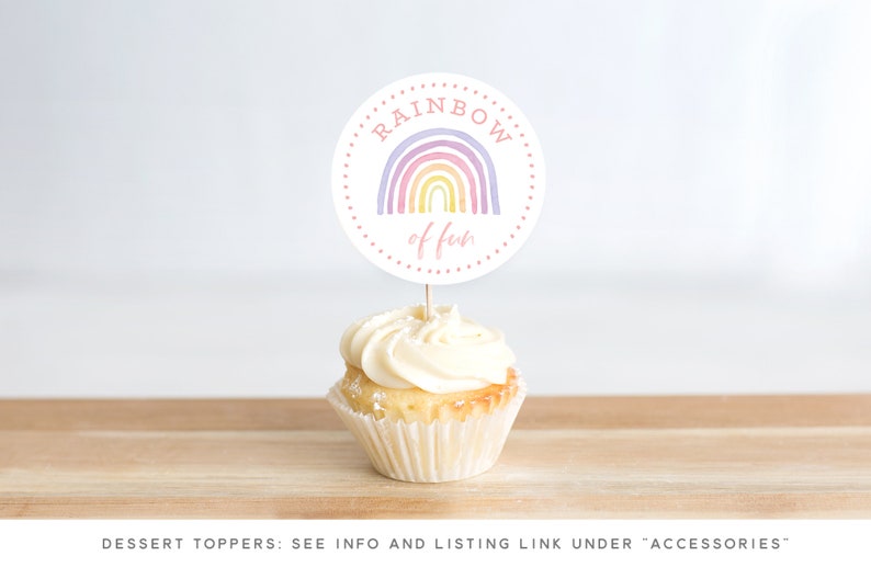 May include: A white cupcake with vanilla frosting topped with a round, white paper cupcake topper that says "RAINBOW of fun" in pink lettering. The topper features a watercolor rainbow with purple, pink, yellow, and orange colors.