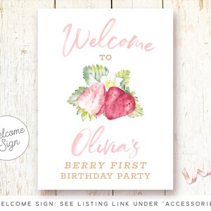 May include: Welcome sign for a first birthday party with a pink and white strawberry design. The sign says "Welcome to Olivia's Berry First Birthday Party".