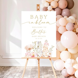 Może przedstawiać: Biały szyld na drewnianym sztaludze ze złotymi literami z napisem "BABY in bloom" i "Welcome to Bella's BABY BRUNCH 08.10.XX". Szyld jest ozdobiony akwarelowymi ilustracjami kwiatowymi i otoczony łukiem z balon&oacute;w.