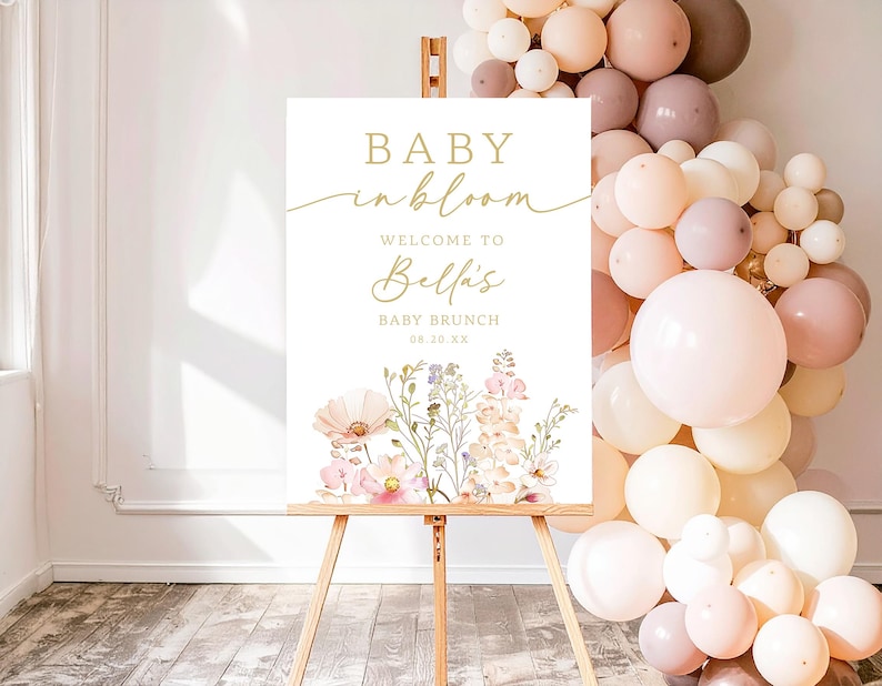 Może przedstawiać: Biały szyld na drewnianym sztaludze ze złotymi literami z napisem "BABY in bloom" i "Welcome to Bella's BABY BRUNCH 08.10.XX". Szyld jest ozdobiony akwarelowymi ilustracjami kwiatowymi i otoczony łukiem z balon&oacute;w.