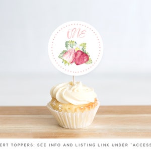May include: A white cupcake with pink frosting and a round, white cake topper with pink polka dots and a watercolor illustration of strawberries. The topper says "One" in pink.