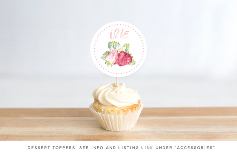 May include: A white cupcake with pink frosting and a round, white cake topper with pink polka dots and a watercolor illustration of strawberries. The topper says "One" in pink.