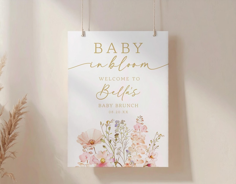 Może przedstawiać: Prostokątny biały znak ze złotym tekstem i ilustracjami kwiatowymi. Tekst brzmi "BABY in bloom" i "WELCOME TO Bella's BABY BRUNCH 08.20.XX". Na dole znaku znajdują się kwiaty w stylu akwareli w odcieniach r&oacute;żu, brzoskwini i fioletu.