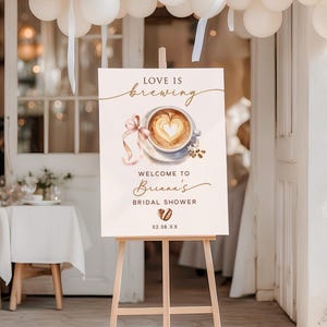 May include: A wooden easel displays a sign with the words "Love is Brewing" above a watercolor image of a coffee cup with a heart-shaped design. The sign welcomes guests to Briana's bridal shower, with the date 02.08.XX.