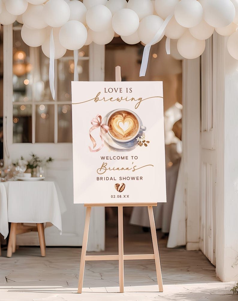 May include: A wooden easel displays a sign with the words "Love is Brewing" above a watercolor image of a coffee cup with a heart-shaped design. The sign welcomes guests to Briana's bridal shower, with the date 02.08.XX.