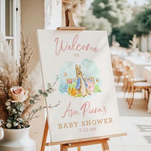 May include: A welcome sign for a baby shower, featuring a watercolor illustration of Peter Rabbit. The sign includes the text "Welcome to Ava Piera's Baby Shower" in pink and gold lettering, displayed on a wooden easel.