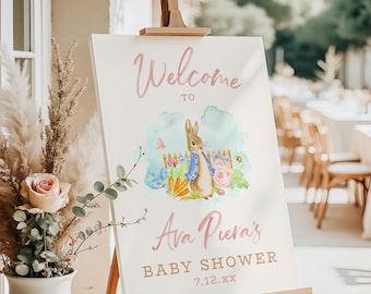 May include: A welcome sign for a baby shower, featuring a watercolor illustration of Peter Rabbit. The sign includes the text "Welcome to Ava Piera's Baby Shower" in pink and gold lettering, displayed on a wooden easel.