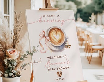 May include: A baby shower welcome sign on an easel. The sign features the text "A Baby Is Brewing" and "Welcome to Brianne's Baby Shower." A watercolor image of a coffee cup with a heart design is also included. The date 09.25.25 is at the bottom.