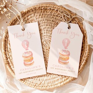 May include: Two pink and white birthday party favor tags with the text "Thank You". Each tag features a watercolor illustration of a stack of donuts topped with a balloon and the number 1. The tags read "CAROLINE'S 1ST BIRTHDAY PARTY 09.25.xx".