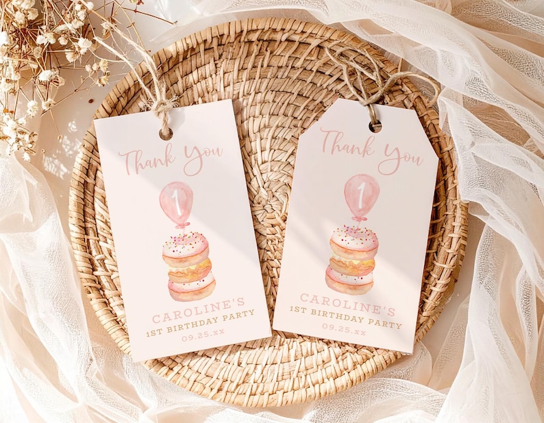 May include: Two pink and white birthday party favor tags with the text "Thank You". Each tag features a watercolor illustration of a stack of donuts topped with a balloon and the number 1. The tags read "CAROLINE'S 1ST BIRTHDAY PARTY 09.25.xx".