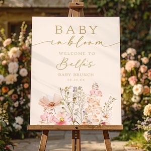 Może przedstawiać: Kremowy znak na drewnianym sztaludze ze złotymi literami z napisem "BABY in bloom" i "WELCOME TO Bella's BABY BRUNCH". Znak jest ozdobiony akwarelowymi ilustracjami kwiatowymi w kolorze r&oacute;żowym, fioletowym i białym.