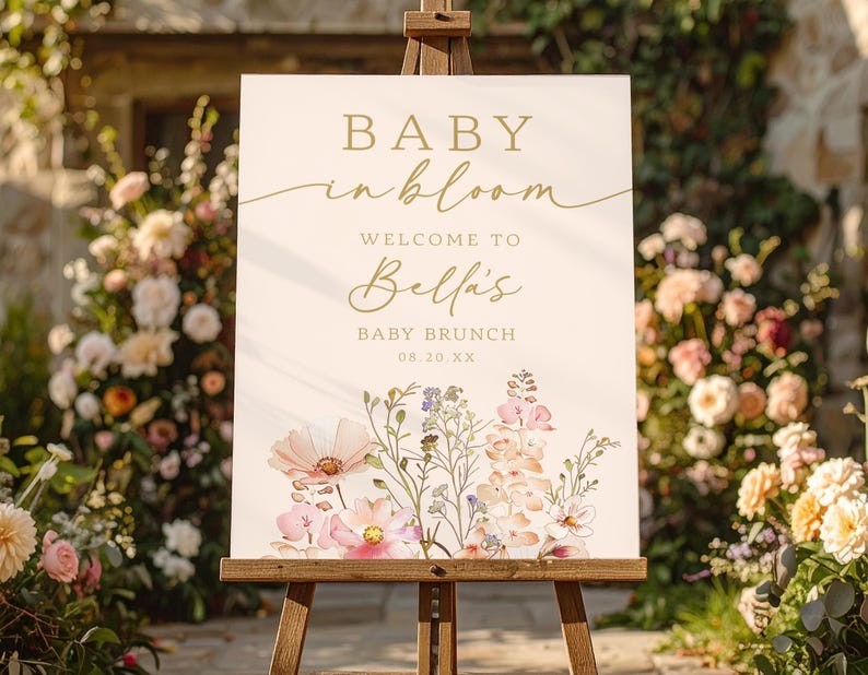 Może przedstawiać: Kremowy znak na drewnianym sztaludze ze złotymi literami z napisem "BABY in bloom" i "WELCOME TO Bella's BABY BRUNCH". Znak jest ozdobiony akwarelowymi ilustracjami kwiatowymi w kolorze r&oacute;żowym, fioletowym i białym.
