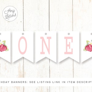 May include: White banner with pink letters spelling "ONE" and strawberry illustrations. The banner is for a first birthday party.