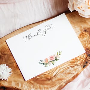 May include: A white card with the words "Thank you" in elegant script, adorned with a watercolor floral arrangement of pink and peach roses and green leaves. The card rests on a wooden surface with a soft pink fabric background.