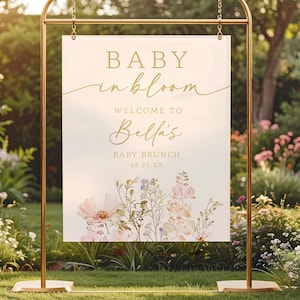 Może przedstawiać: Złota metalowa rama trzyma znak z napisem "BABY in bloom" i "Welcome to Bella's BABY BRUNCH 08.20.XX". Znak zawiera kwiatowe ilustracje w miękkich r&oacute;żach i fioletach na jasnym tle.
