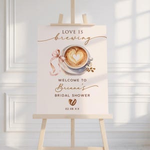 May include: A cream-colored sign on a wooden easel with the words "Love is Brewing" in elegant script. A watercolor image of a coffee cup with a heart-shaped design is featured. The sign welcomes guests to Briana's Bridal Shower.