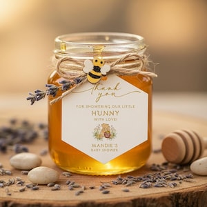 May include: A small glass jar filled with golden honey, adorned with a twine bow, a wooden bee, and a label that reads "Thank you for showering our little Hunny with love!" A wooden honey dipper and lavender sprigs are nearby.
