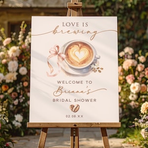 May include: A white sign on a wooden easel with the text "LOVE IS brewing" and "WELCOME TO Briana's BRIDAL SHOWER". The sign features a watercolor illustration of a coffee cup with a heart-shaped design in the foam, and a pink ribbon.