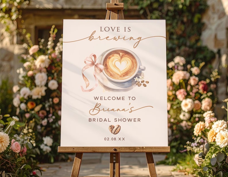 May include: A white sign on a wooden easel with the text "LOVE IS brewing" and "WELCOME TO Briana's BRIDAL SHOWER". The sign features a watercolor illustration of a coffee cup with a heart-shaped design in the foam, and a pink ribbon.