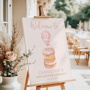 May include: A pink sign on a wooden easel with the words "Welcome to" and "Caroline's 2nd Birthday Party." The sign features a watercolor illustration of a balloon and a stack of donuts.