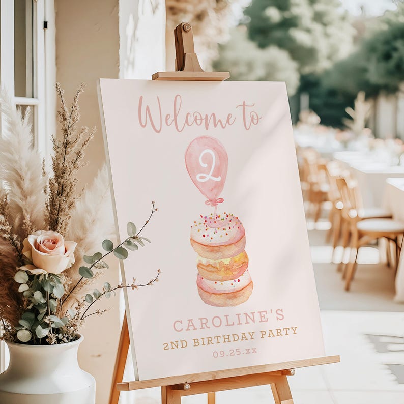 May include: A pink sign on a wooden easel with the words "Welcome to" and "Caroline's 2nd Birthday Party." The sign features a watercolor illustration of a balloon and a stack of donuts.