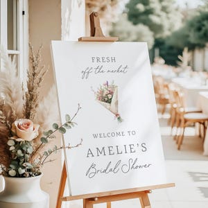 May include: A white sign on a wooden easel with the text "FRESH off the market" and "WELCOME TO AMELIE'S Bridal Shower." A watercolor illustration of a bouquet of flowers is also on the sign. A vase of flowers is in the foreground.
