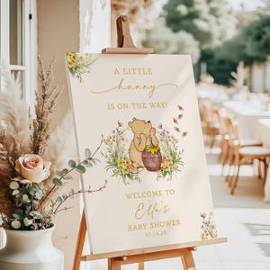 May include: A cream-colored baby shower welcome sign on a wooden easel. The sign features a Winnie the Pooh illustration with the text "A Little Hunny Is On The Way!" and "Welcome to Elli's Baby Shower 03.14.2X."