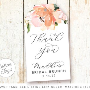 May include: A white thank you tag with a floral design in shades of pink and orange. The text "Thank you" is written in cursive script. The text "Maddie's Bridal Brunch 5.14.22" is printed below the floral design.