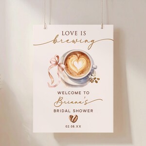 May include: A rectangular sign with the words "Love is Brewing" above a watercolor image of a coffee cup with a heart-shaped design in the foam. The sign reads "Welcome to Briana's Bridal Shower" with a date below.