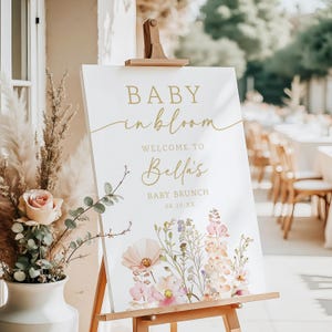 Może przedstawiać: Biały znak na drewnianym sztaludze z napisem "BABY in bloom" złotym skryptem. Znak wita gości na Baby Brunch Belli, z datą poniżej. Kwiatowe ilustracje w kolorze r&oacute;żowym, brzoskwiniowym i fioletowym zdobią d&oacute;ł.